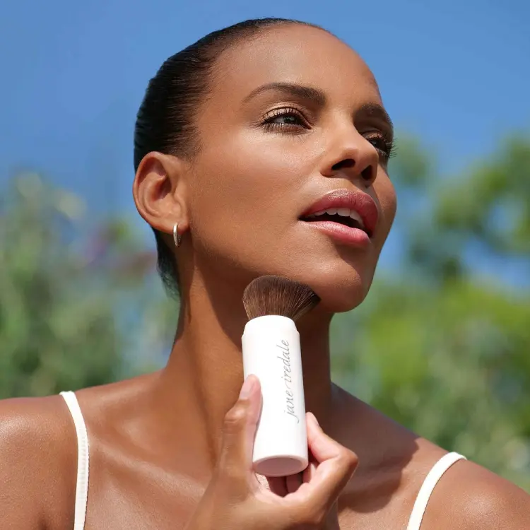 Na zdjęciu widać kobietę aplikującą puder za pomocą pędzla jane iredale Powder-Me SPF. Produkt ma formę wygodnego aplikatora z pędzlem i jest używany na zewnątrz, w słoneczny dzień, co podkreśla jego funkcję ochrony przeciwsłonecznej (SPF).