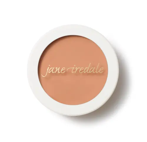 Na zdjęciu jest korektor w kremie marki Jane Iredale Enlighten Concealer, zamknięty w okrągłym opakowaniu z białą pokrywką. Na wieczku widnieje eleganckie złote logo "jane iredale". Produkt ma ciepły, beżowo-brzoskwiniowy odcień.
