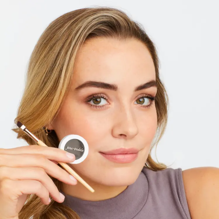 Na zdjęciu widzimy kobietę w naturalnym makijażu, która prezentuje jane iredale PureBrow Brow Powder. W jednej ręce trzyma puder do brwi i drugiej pędzel. Jej brwi są starannie podkreślone, są pełniejsze i mają miękko zarysowany kształt.