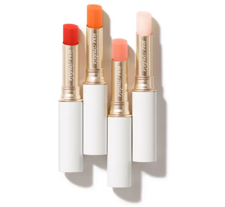 Na zdjęciu widoczne są cztery pomadki z kolekcji Just Kissed Lip and Cheek Stain marki Jane Iredale. Produkty umieszczone są w eleganckich, białych opakowaniach ze złotym wykończeniem.  Każda pomadka ma inny odcień