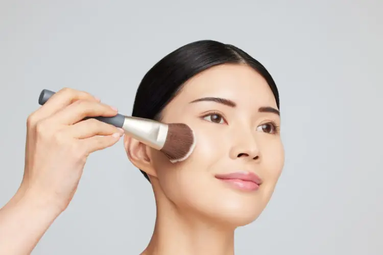 Na zdjęciu widzimy kobietę, której twarz jest pokrywana pudrem przy użyciu pędzla Jane Iredale Multiuse. Pędzel ma elegancki wygląd — jasne włosie przechodzące w ciemniejsze u nasady, złotą skuwkę oraz szarą rączkę z logotypem.