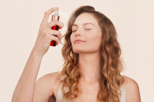 Na zdjęciu kobieta spryskuje twarz nawilżającą mgiełką Pommist Hydration Spray od Jane Iredale. Ma zamknięte oczy i zrelaksowany wyraz twarzy. Produkt widoczny w jej dłoni to czerwona butelka z białym atomizerem – idealny dla każdego rodzaju skóry.