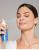 Na zdjęciu kobieta spryskuje twarz nawilżającą mgiełką D₂O™ Hydration Spray od Jane Iredale. Ma zamknięte oczy i zrelaksowany wyraz twarzy. Produkt widoczny w jej dłoni to niebieska butelka z białym atomizerem – idealny do skóry suchej.