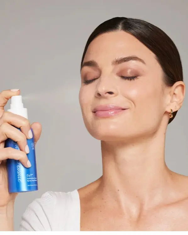 Na zdjęciu kobieta spryskuje twarz nawilżającą mgiełką D₂O™ Hydration Spray od Jane Iredale. Ma zamknięte oczy i zrelaksowany wyraz twarzy. Produkt widoczny w jej dłoni to niebieska butelka z białym atomizerem – idealny do skóry suchej.