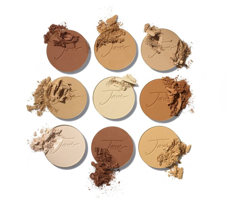 Na zdjęciu jest dziewięć okrągłych pudrów prasowanych Jane Iredale w różnych odcieniach – od bardzo jasnych po ciemne, idealnych do dopasowania do różnych tonacji skóry. Część z nich jest lekko pokruszona, co podkreśla konsystencję produktu.