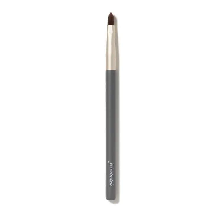 Na zdjęciu jest cienki pędzelek do makijażu jane iredale. Ma smukły kształt, szarą rączkę z logo marki i złotą skuwkę. Włosie pędzelka jest krótkie, ścięte w szpic, co sprawia, że idealnie nadaje się do precyzyjnej aplikacji produktów.