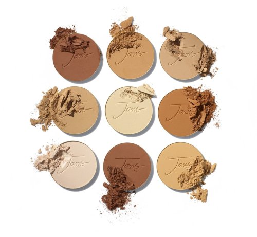Na obrázku je vidět devět kulatých lisovaných pudrů Jane Iredale v různých odstínech – od velmi světlých po tmavé, ideálních pro přizpůsobení různým tónům pleti. Některé z nich jsou lehce rozdrcené, což zdůrazňuje texturu produktu.