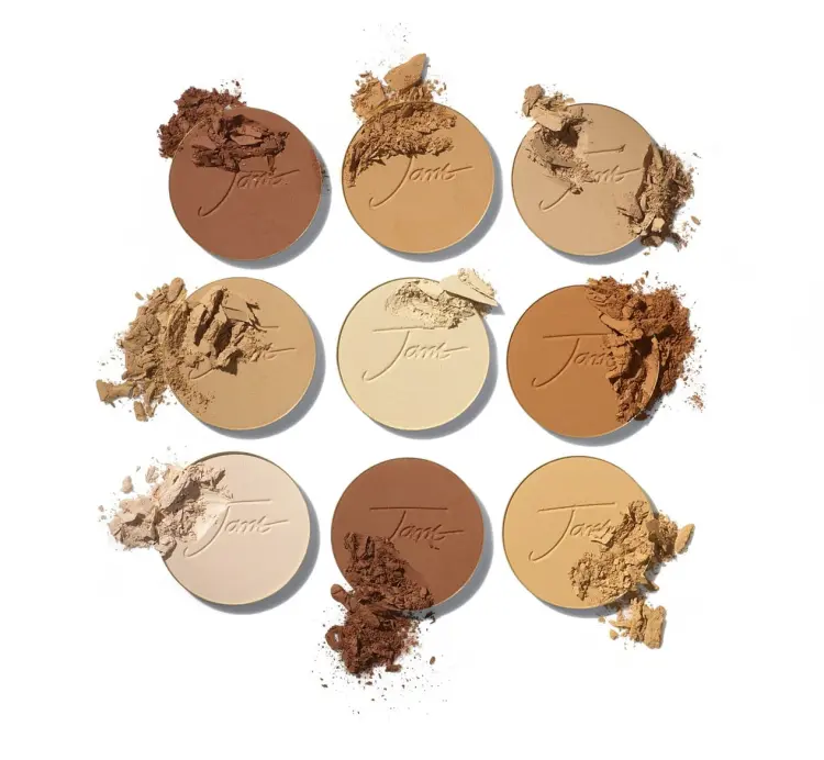 Na obrázku je vidět devět kulatých lisovaných pudrů Jane Iredale v různých odstínech – od velmi světlých po tmavé, ideálních pro přizpůsobení různým tónům pleti. Některé z nich jsou lehce rozdrcené, což zdůrazňuje texturu produktu.
