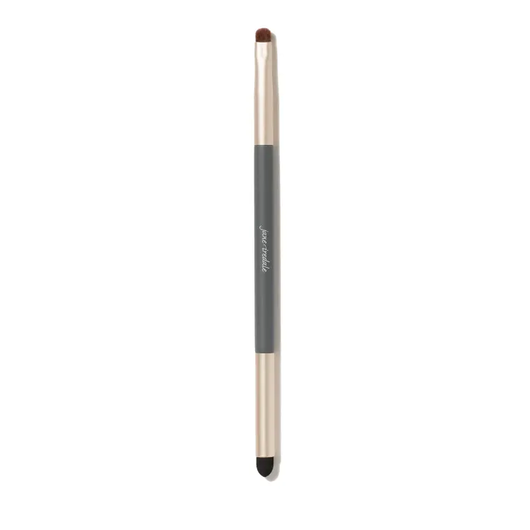 Zdjęcie przedstawia dwustronny pędzelek do makijażu jane iredale na białym tle. Ma szary trzonek z logo marki i złote skuwki. Z jednej strony znajduje się mały, zaokrąglony pędzelek z brązowym włosiem, a z drugiej czarny aplikator gąbkowy.