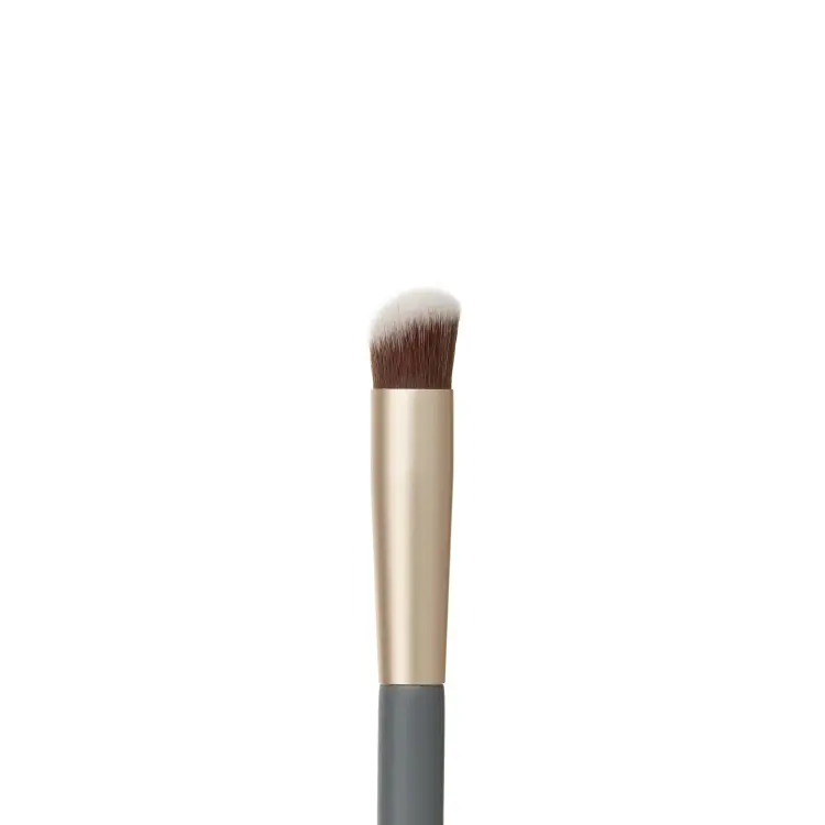 Zdjęcie przedstawia zbliżenie końcówki pędzelka do korektora jane iredale Concealer Brush na białym tle. Widoczne jest gęste, lekko skośne włosie w kolorze brązowym z białymi końcówkami, osadzone w złotej skuwce połączonej z szarym trzonkiem.