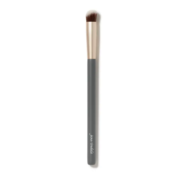 Zdjęcie przedstawia pędzelek do korektora jane iredale na białym tle. Ma długi, szary trzonek z białym logo marki, złotą skuwkę oraz krótkie, gęste, lekko zaokrąglone włosie w kolorze brązowym z białymi końcówkami.