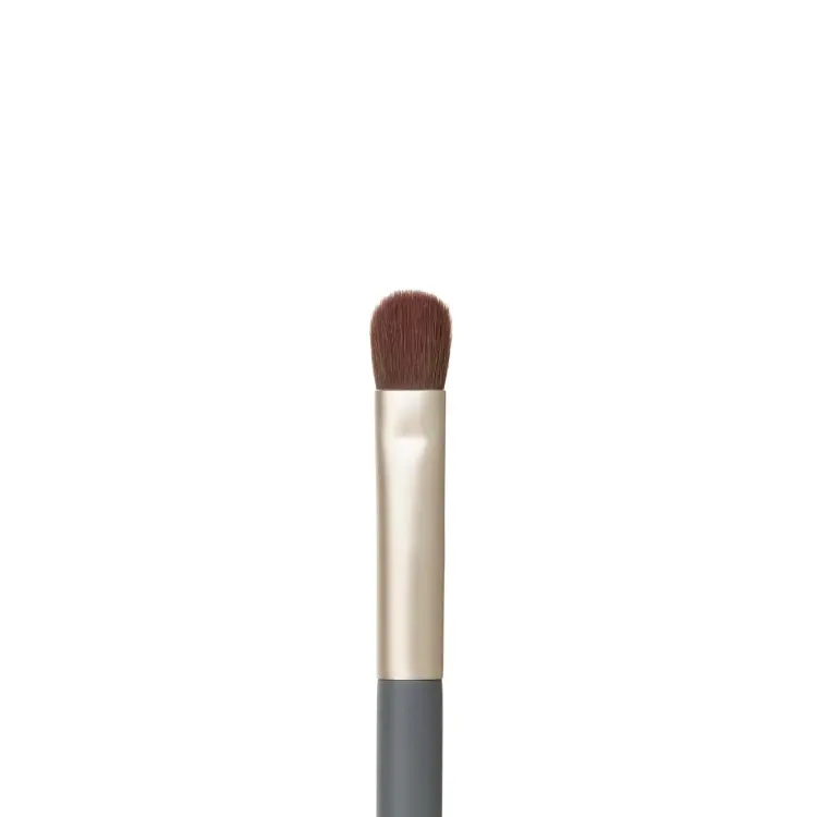 Zdjęcie przedstawia zbliżenie końcówki pędzelka do makijażu oczu jane iredale Flat Eye Brush na białym tle. Widoczne jest krótkie, gęste, płasko ułożone brązowe włosie o zaokrąglonej końcówce, osadzone w złotej skuwce połączonej z szarym trzonkiem.
