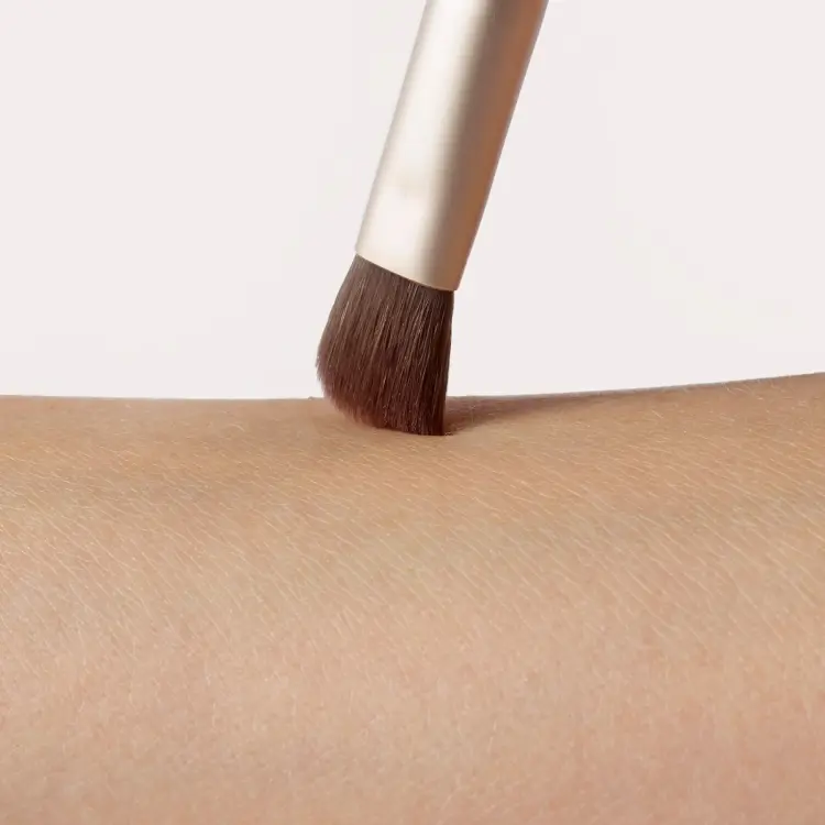 Zdjęcie przedstawia zbliżenie pędzelka jane iredale Flat Eye Brush podczas kontaktu ze skórą. Gęste, płasko ułożone brązowe włosie delikatnie dotyka przedramienia, ukazując kształt i precyzję końcówki. Widoczna jest również złota skuwka pędzelka.