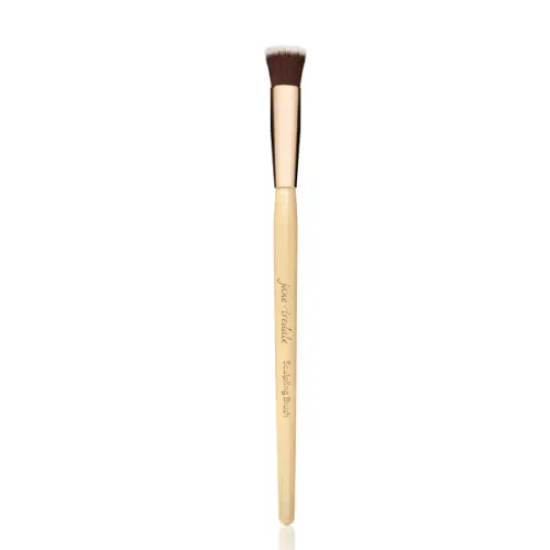 Na zdjęciu Sculpting Brush marki Jane Iredale, krótki, płaski, ścięty pod kątem, do konturowania  Rączka drewniana z jasnego drewna, złota skuwka, nadrukowana nazwa marki i pędzla  Włosie syntetyczne, zbite, w kolorze ciemnobrązowym z jasnymi końcówkami