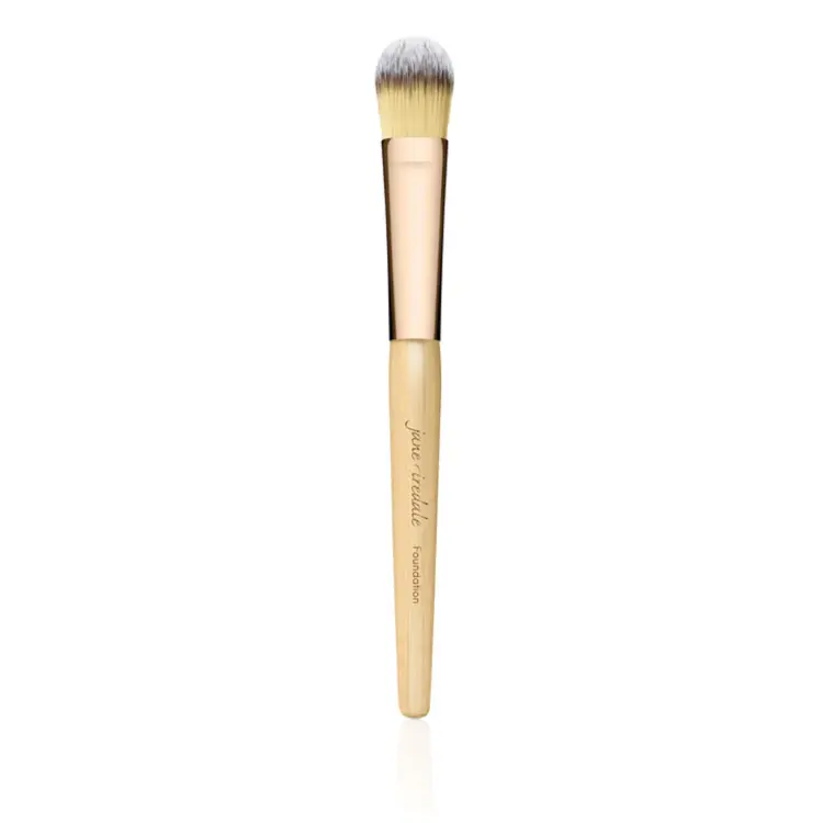 Na zdjęciu znajduje się pędzel do makijażu marki Jane Iredale. Ma elegancki design – drewniany trzonek w naturalnym kolorze z złotą skuwką i gęste, miękkie włosie w odcieniach żółci i bieli.